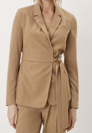 Beżowy garnitur na miarę z marynarką z klapami z nacięciem, ozdobioną bocznym paskiem do wiązania, noszony z dopasowanymi spodniami z wysokim stanem, pokazany na osobie od szyi do połowy uda.