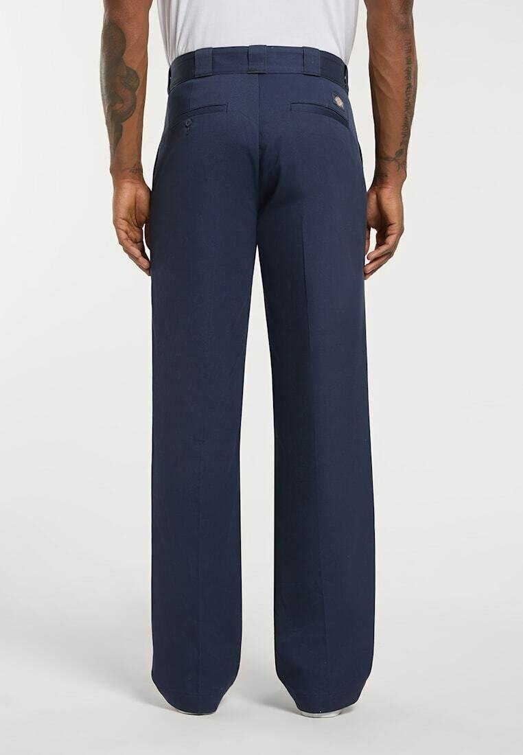 Man wearing navy blue wide-leg pants, standing with arms relaxed, visible tattoos on forearms, plain white background.