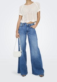 Bílý pletený crop top s krátkými rukávy, doplněný širokými modrými džíny. Drží malou bílou kabelku. Hladké textury, ležérní design.