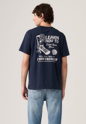 Homme portant un t-shirt bleu marine avec une impression blanche d’un téléphone à clapet, une fleur et un texte sur le fait de laisser tomber les téléphones pour des activités en plein air.