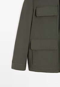 Giacca verde oliva realizzata in tessuto liscio, con chiusura a zip, due tasche con pattina frontali e un design semplice con dettagli trapuntati.