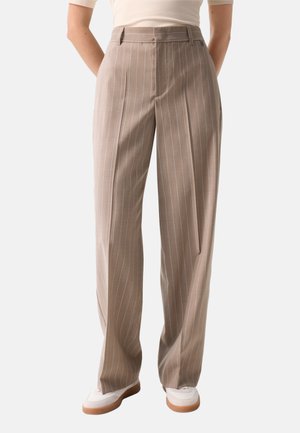 Beige Nadelstreifen-Hosen mit weitem Bein, getragen mit weißen Schuhen und einem hellbeigen Oberteil vor einfarbigem Hintergrund.
