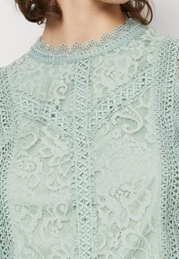 Haut en dentelle bleu clair avec motifs floraux, broderie délicate et encolure en dents de scie avec des accents décoratifs.