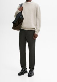 Pull beige en maille, pantalons foncés et chaussures noires. Tient un sac marron avec un objet gris à l'intérieur. Design simple, texture douce.