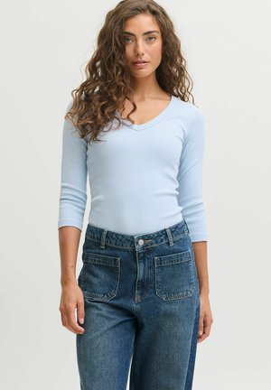 Femme aux cheveux bouclés portant un haut moulant bleu clair à manches trois-quarts et un jean bleu taille haute avec de grandes poches avant.