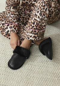 Čierne papuče s mäkkým čiernym kožušinovým lemom a textúrovaným trblietavým povrchom, nosené s pyžamami s leopardím vzorom na svetlom tkanom koberci.