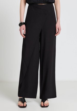 Donna che indossa pantaloni neri a vita alta e gamba larga con un pannello frontale asimmetrico, abbinati a sandali neri con punta aperta e un bracciale a polsino nero.