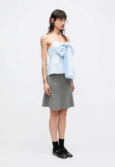 Svetlomodrý top na ramienka s veľkým mašľovým detailom, v kombinácii so sivou, kolenovou rozšírenou sukňou. Modelka má na sebe čierne ponožky a topánky.