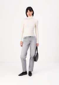 Un col roulé blanc à côtes, un jean gris à jambe droite et des chaussures noires à clous. Tenant un petit sac rond noir avec une surface texturée.