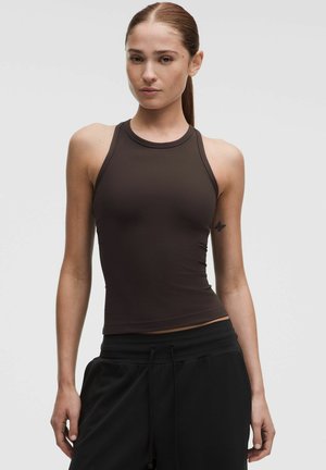 Junge Frau mit langem braunem Pferdeschwanz, trägt ein ärmelloses dunkelbraunes Top und schwarze Hose, steht vor einem schlichten Hintergrund.