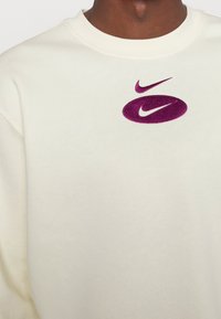 Krémová mikina s fialovým vyšívaným logem Nike. Látka má hladkou texturu, obléká se přes hlavu a má spuštěná ramena.