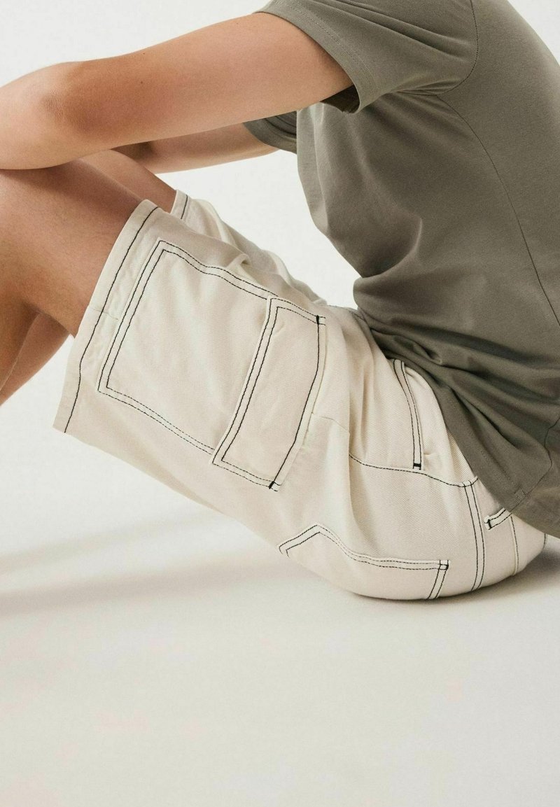 Shorts beige avec coutures visibles et deux grandes poches, associés à un t-shirt gris. Le tissu semble lisse et léger.