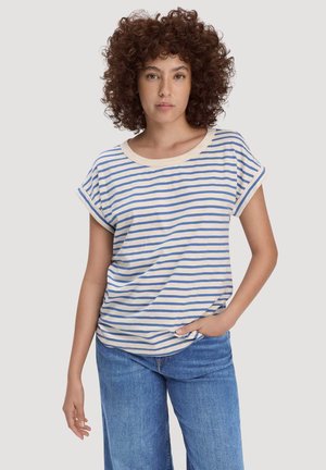 Woman with curly hair wearing a cream and blue striped short-sleeve shirt and blue jeans, standing with one hand in pocket against a plain background.