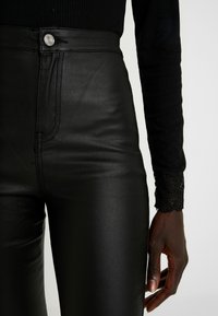 Svarta läderbyxor med hög midja och en slät yta, med metalldynan och kontrasterande spetsdetaljer på ärmar av svart tyg.