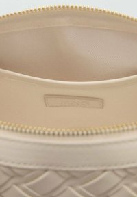 Beige zipperpussi, joka on valmistettu sileästä materiaalista, jossa on kudottu kuvio ulkopuolella ja pehmeä vuori, jossa on pieni logo-tarra.