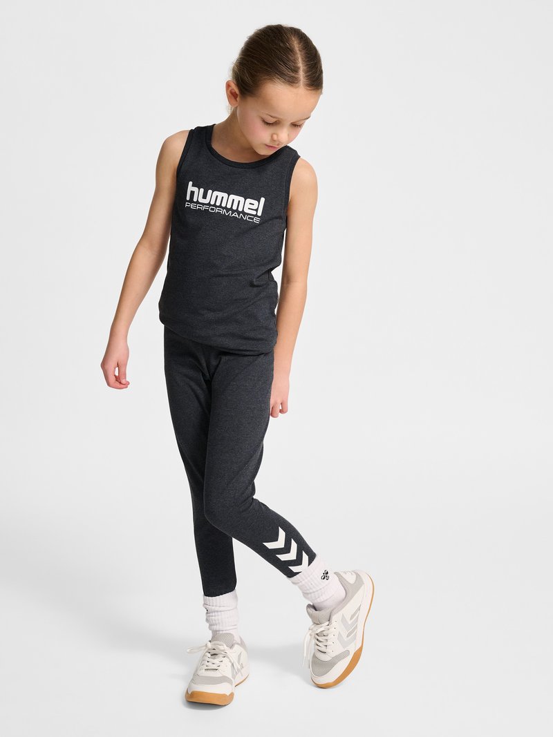 Junges Mädchen trägt ein dunkles sportliches Tanktop und Leggings mit weißem Chevron-Logo, weiße Socken und hellgraue Turnschuhe, schaut nach unten.