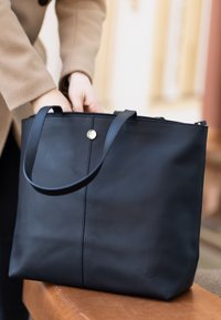 Zwarte leren tote bag met een gladde textuur, voorzien van twee handvatten, een ritssluiting aan de bovenkant en een gouden drukknoopdetail aan de voorkant.