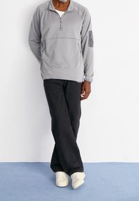 Grå fleece-pullov med halv dragkedja, två framfickor och en sidoficka på ärmen; tillsammans med svarta jeans och krämfärgade sneakers.