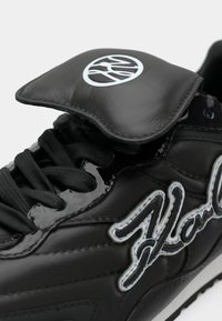 Crna tenisica s sjajnim završetkom, vezicama i podstavljenim jezičkom s okruglim logotipom. Bijeli šavovi ističu detalje dizajna.