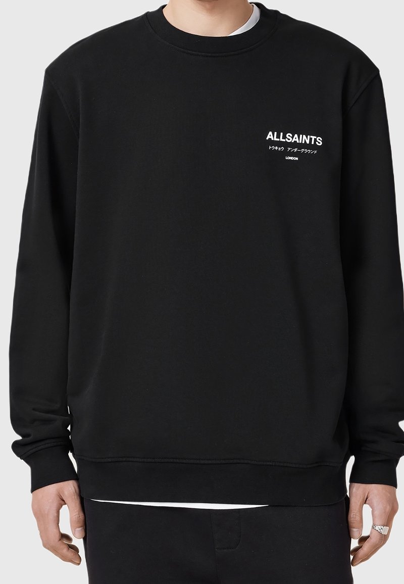 Sweat-shirt noir à manches longues et col rond avec un petit texte blanc « ALLSAINTS » et du texte japonais sur le côté gauche de la poitrine, porté par une personne debout.