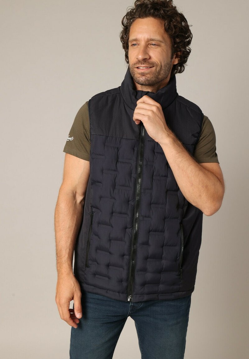 Gilet rembourré bleu marine avec un col haut, une fermeture éclair et une texture matelassée. Présente des accents en maille noire et deux poches latérales zippées.
