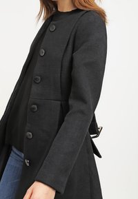 Schwarzer Wollmantel mit rundem Ausschnitt, langen Ärmeln, sechs Knopfverschlüssen, seitlichen Taschen und einem taillierten Rückendetail.