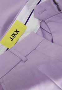 Šviesiai violetinės kelnės su lygiu paviršiumi, turinčios geltoną etiketę su užrašu "JJXX" ir baltą pamušalą su violetiniu akcentu.