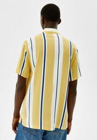 Camisa de botones de manga corta en amarillo con rayas verticales en blanco y azul marino, que presenta un cuello clásico y un corte relajado.