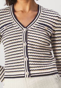 Kobieta nosząca sweter w paski z perełkowymi guzikami i dekoltem w kształcie litery V, zestawiony z jasnymi, teksturowanymi spodniami.