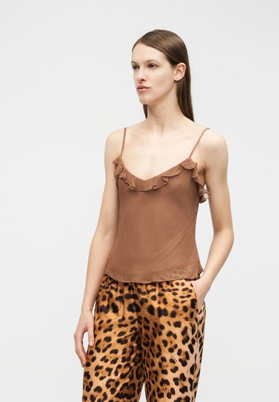 Γυναίκα με ίσια καστανά μαλλιά, που φοράει καφέ τοπ με ριχτά τιραντάκια και παντελόνι με λεοπάρ σχέδιο, στέκεται με το ένα χέρι στην τσέπη.