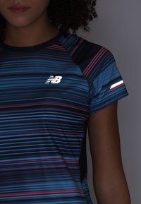 Camiseta deportiva de manga corta en azul marino con rayas horizontales en diferentes tonos de azul y rojo, que presenta un logo blanco en el pecho.