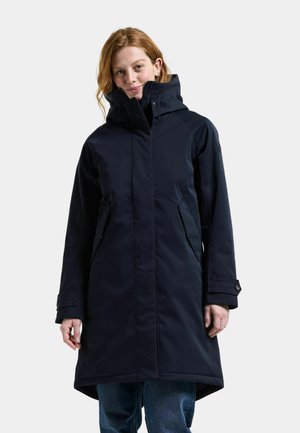 Kvinde iført en lang sort hættejakke med lynlås og lommer, kombineret med blå jeans, stående mod en ensfarvet lys baggrund.