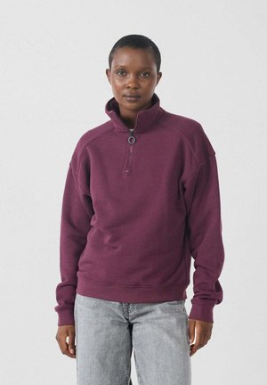Pullover in pile bordeaux con zip a un quarto, maniche raglan e polsini a coste. Indossato con jeans grigio chiaro su uno sfondo neutro.