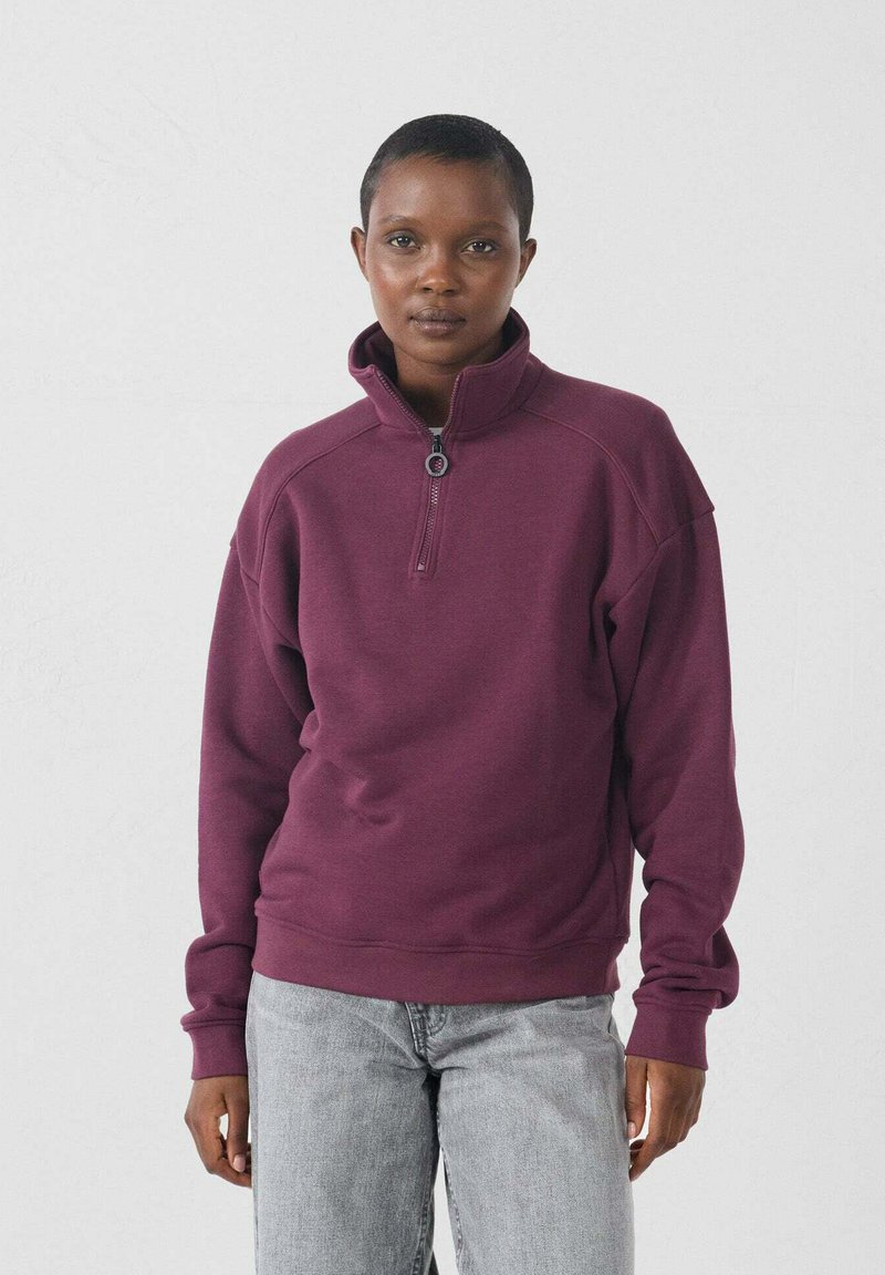 Pullover in pile bordeaux con zip a un quarto, maniche raglan e polsini a coste. Indossato con jeans grigio chiaro su uno sfondo neutro.