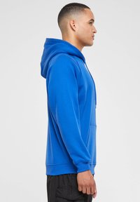 Sudadera de algodón azul con bolsillo frontal y capucha ajustable, con puños y dobladillo acanalados, exhibida en un modelo masculino en vista lateral.
