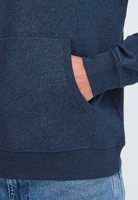 Mano dentro della tasca anteriore di una felpa blu navy indossata con jeans in denim azzurro su uno sfondo neutro.