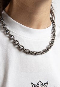Sølvkjede laget av store sammenkoblede lenker, vist over en hvit bomulls T-skjorte med subtil broderi.