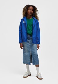 Blå letvægtsjakke, grøn t-shirt, denim midi-nederdel med frontslids. Cremefarvede sneakers med grå detaljer; hvide sokker synlige.