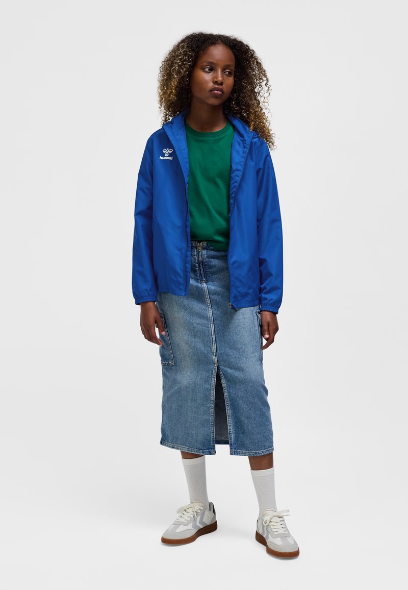 Blå letvægtsjakke, grøn t-shirt, denim midi-nederdel med frontslids. Cremefarvede sneakers med grå detaljer; hvide sokker synlige.