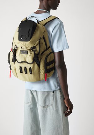 Zaino khaki con spallacci e dettagli neri. Presenta diverse tasche frontali, un retro imbottito e zip rosse; realizzato in materiale testurizzato.