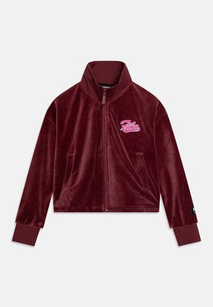 Bordeauxrode jas met rits van zachte fluweelstof met geribde kraag en manchetten. Heeft een geborduurd roze logo op de linkerborst en twee zijzakken.