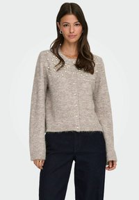 Hellbeige Strickjacke mit rundem Ausschnitt, verziert mit Perlen am Brustbereich und einem verkürzten Saum, getragen über dunklen Jeans.