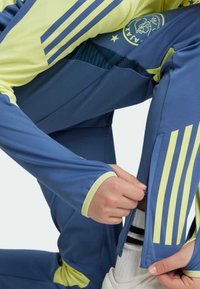 Survêtement bleu et jaune à manches longues et pantalons zippés. Présente trois bandes verticales sur les manches et un emblème d'équipe sur la poitrine.