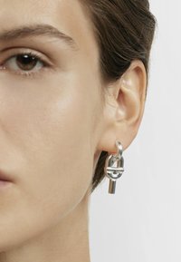 Pendiente de eslabones de cadena de plata que presenta un diseño de cerradura y un acento colgante. Textura lisa y brillante con una forma geométrica moderna.