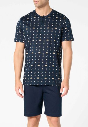 Uomo che indossa una maglia henley blu navy a maniche corte con piccoli motivi di camper e palme, abbinata a pantaloncini blu navy semplici.