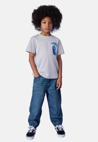 Graues T-Shirt mit blauem Grafikdesign auf der Brust, kombiniert mit locker sitzenden blauen Jeans und schwarzen Turnschuhen. Das Kind steht mit Händen in den Taschen.