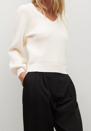 Kobieta w białym dzianinowym swetrze z dekoltem w serek, z długimi rękawami, oraz wysokim stanem czarnych plisowanych spodni, trzymająca ręce w kieszeniach.