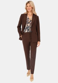Brązowy garnitur dopasowany z blazerem i spodniami prostymi. Wewnętrzna bluzka ma kolorowy kwiatowy wzór. Beżowe szpilki uzupełniają stylizację.