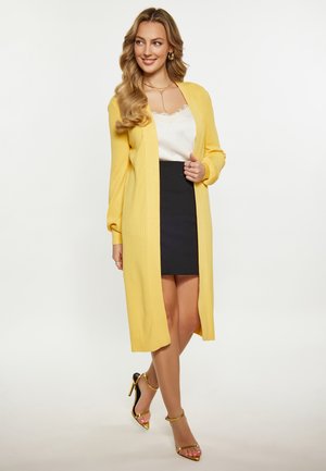 Vrouw glimlacht, draagt een lange gele cardigan over een wit kanten topje en een zwarte rok, met gouden sandalen met hoge hakken en sieraden.