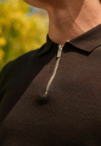 Mies, jolla on päällään tummanruskea, teksturoitu poolopaita, jossa on hopeanvärinen etuvetoketju osittain suljettuna, ja hän seisoo hämärässä ulkona taustalla.